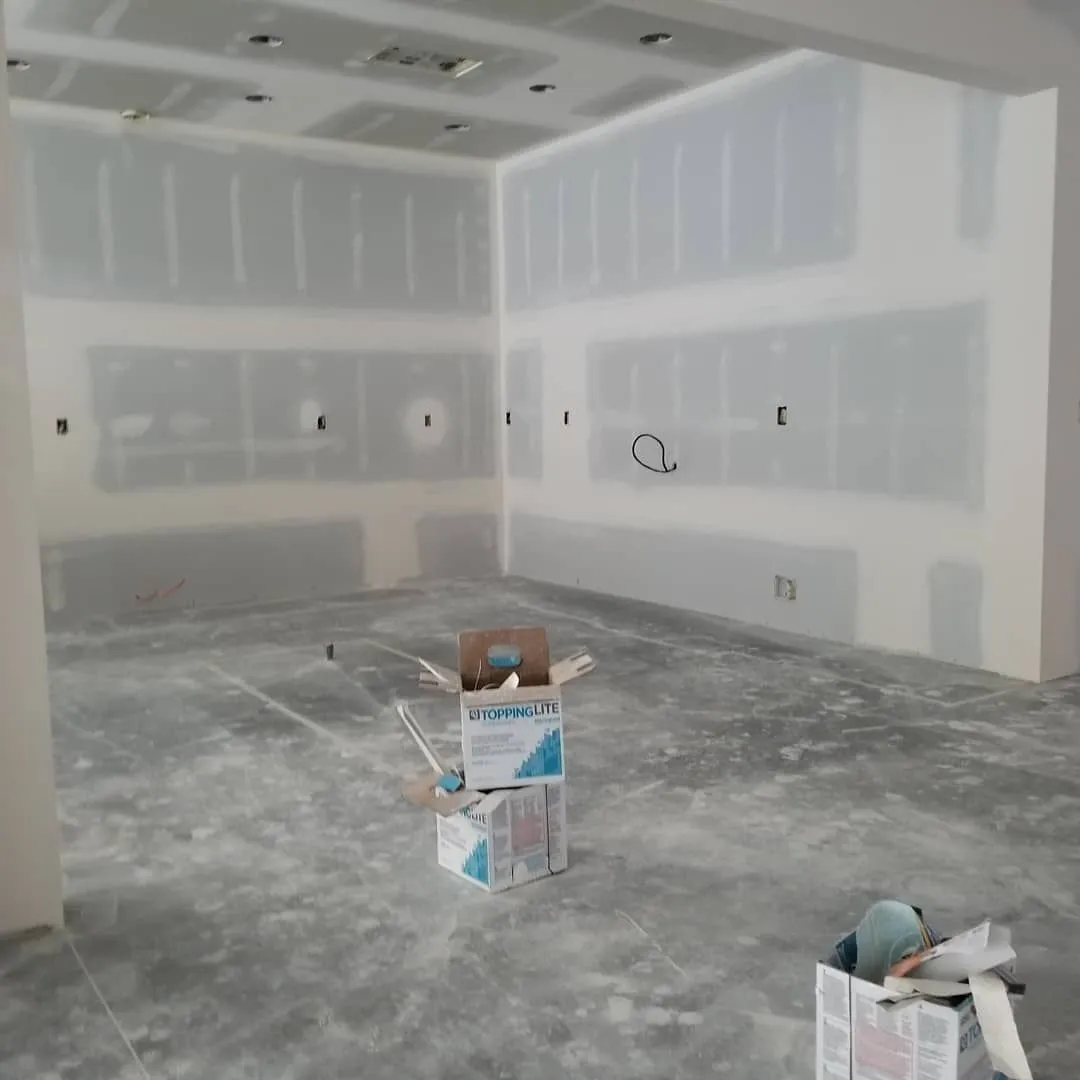Unfinished drywall room with white joint compound, paint supplies on the floor, and exposed electrical outlets.