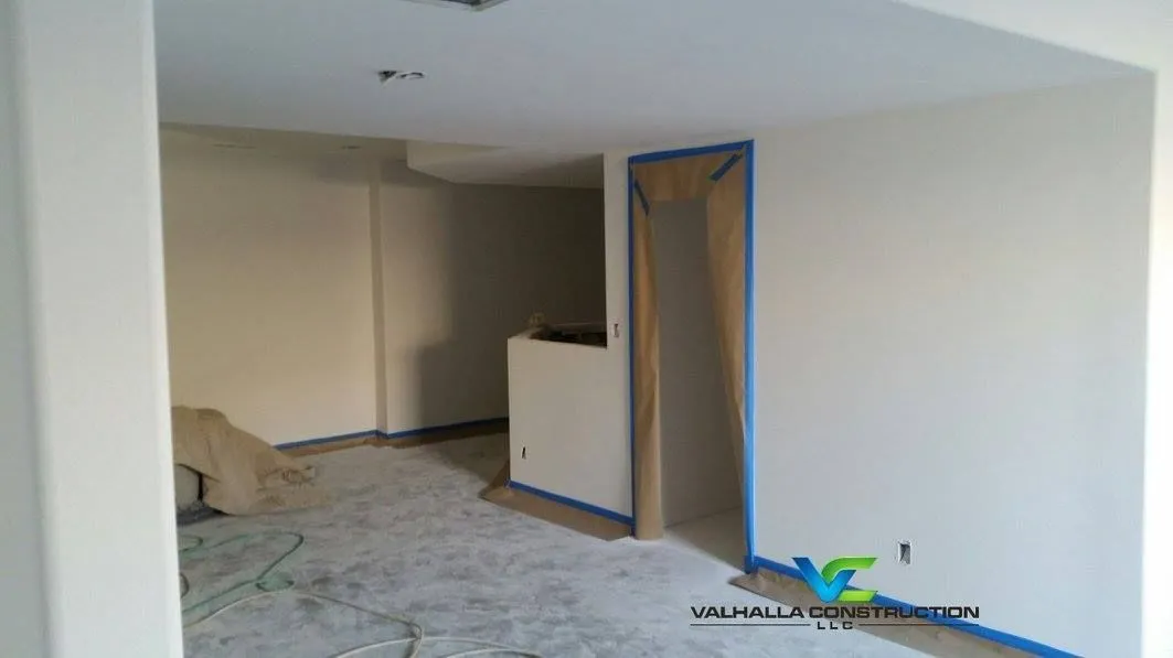 Interior room under renovation, light-colored walls, open doorway, blue tape, unfinished floor.