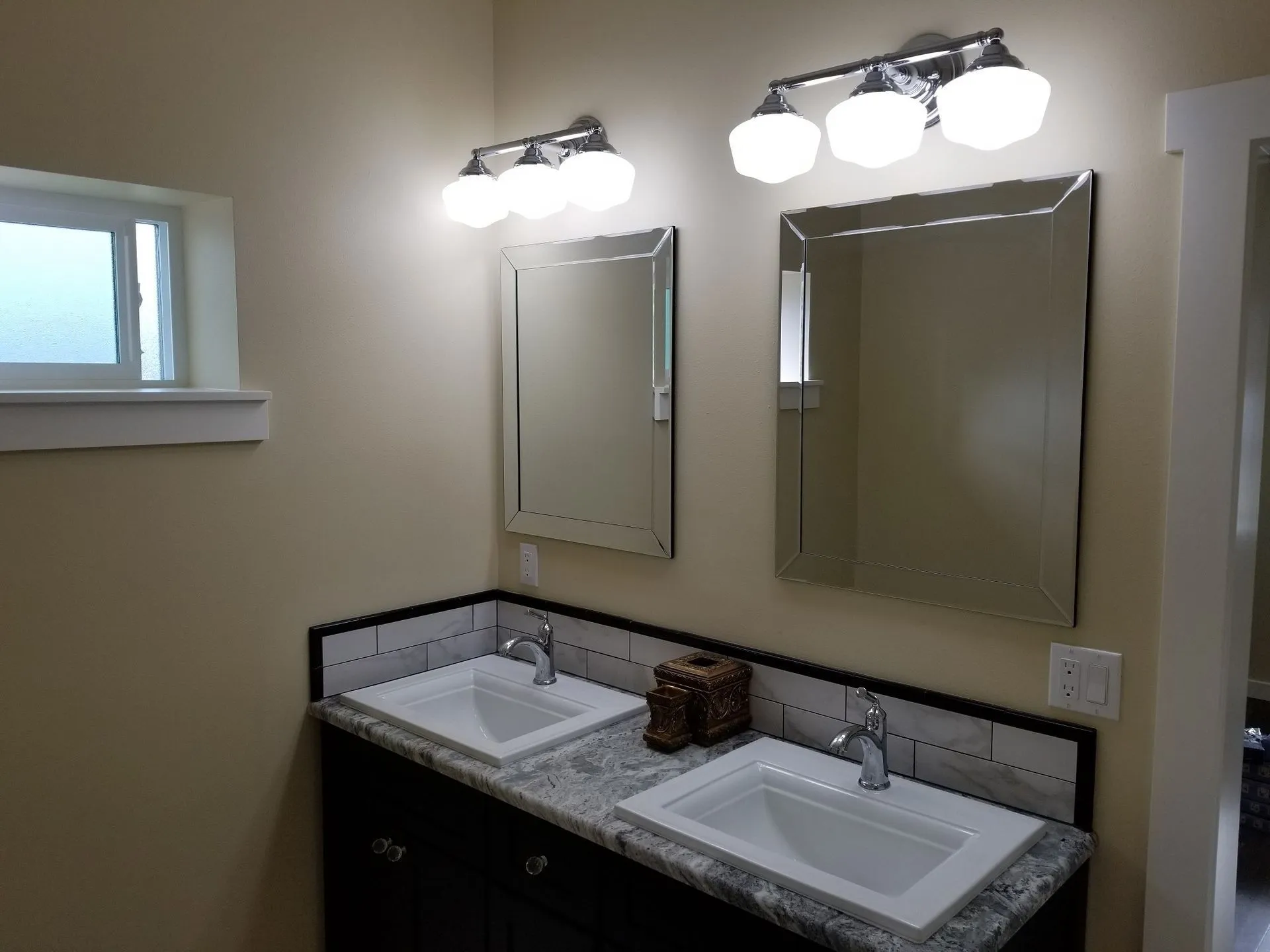 Bathroom with two sinks, mirrors, and overhead lighting fixtures. Cream walls, dark vanity, and window.