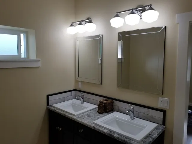 Bathroom with two sinks, mirrors, and overhead lighting fixtures. Cream walls, dark vanity, and window.