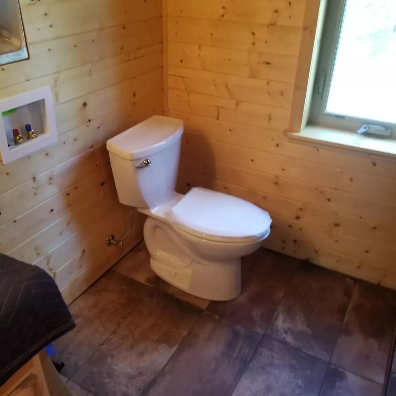 Toilet in a small bathroom with wood-paneled walls, a window, and tiled floor.