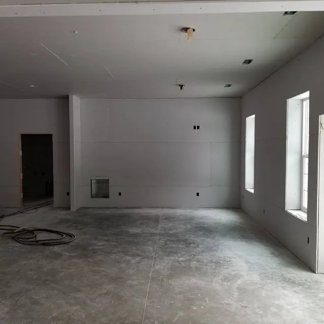 Interior room under construction with gray concrete floor, white walls, and two windows.