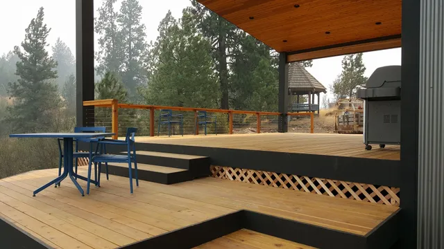 Wooden deck with blue table/chairs, leading to a covered patio. Grill, trees in background.