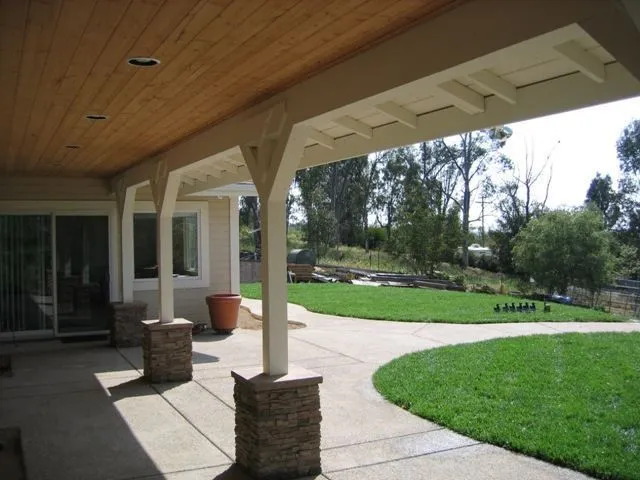 Covered patio with stone pillars, concrete flooring, and a backyard with green grass and trees.