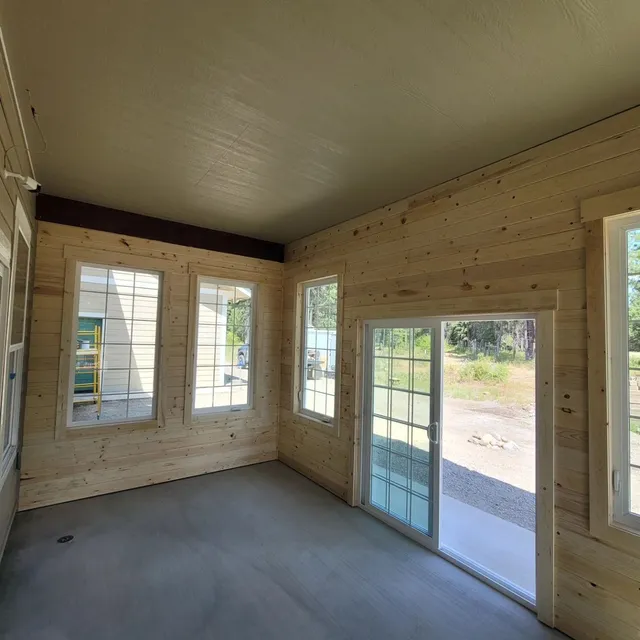 An unfinished room with wooden paneling, windows, and sliding glass door leading to a patio.