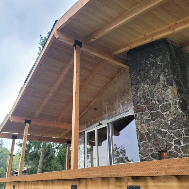 Wooden porch overhang supported by columns next to a stone chimney and window.