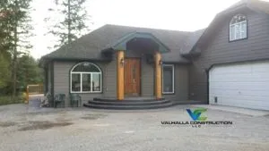 Beige house with wooden door and pillars, gray roof, curved entryway, and gravel driveway.