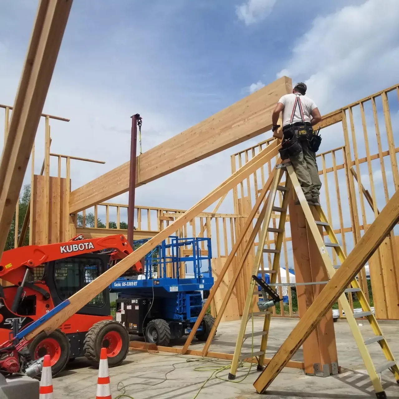Construction worker on ladder, placing wooden beam. Red Kubota, blue lift, building frame. Sunny day.