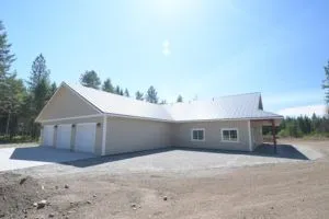 Tan, modern building with three-car garage and attached single-story section with porch. Gravel driveway, bright sky.