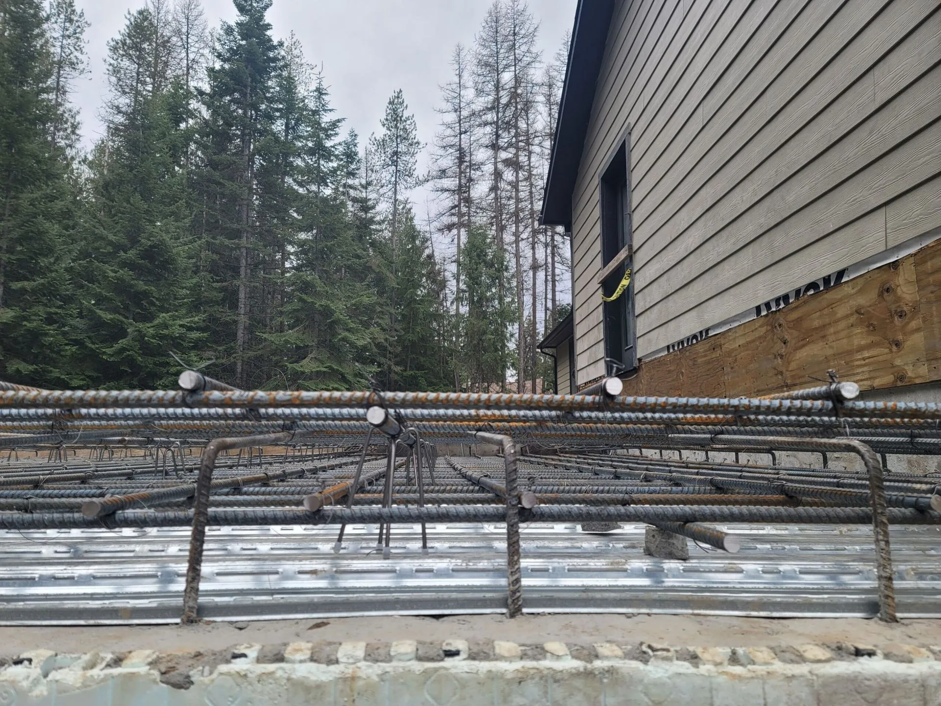 Rebar framework prepared on corrugated metal for a concrete foundation, next to a wood-sided building.