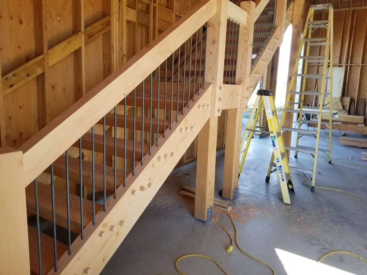Wooden staircase under construction, with metal spindles. A-frame ladder visible.