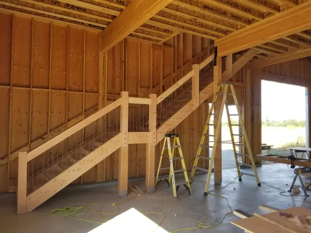 Wooden staircase under construction inside a building. Two ladders support the structure.