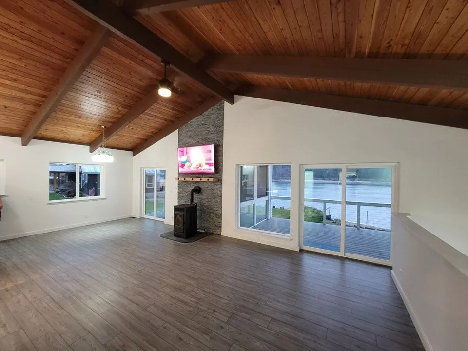 Spacious living room with vaulted wood ceiling, fireplace, large windows overlooking a body of water.